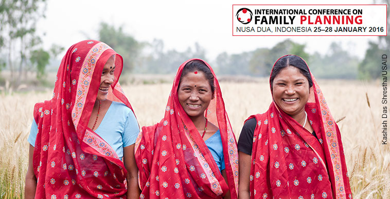 Three women dressed in red saris stand in a field with the logo for the International Conference on Family Planning, Nusa Dua, Indonesia January 25-28, 2016. Photograph by Kashish Das Shrestha/USAID.