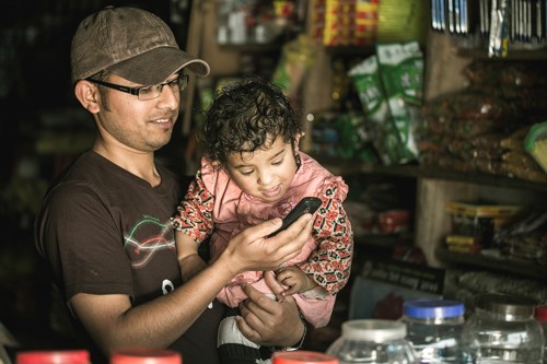 A young child looks on as his father, a small shop owner, uses his low-cost cell phone to pay his suppliers. 
