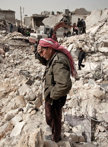 A Syrian man reacts while standing on the rubble of his house, while others look for survivors and bodies in the Tariq al-Bab di