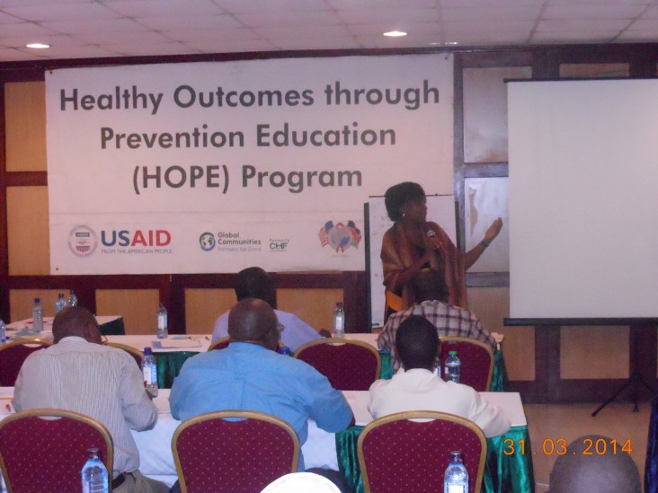 A Kenyan woman addresses an audience while standing in front of a banner that reads Health Outcomes Through Prevention Education