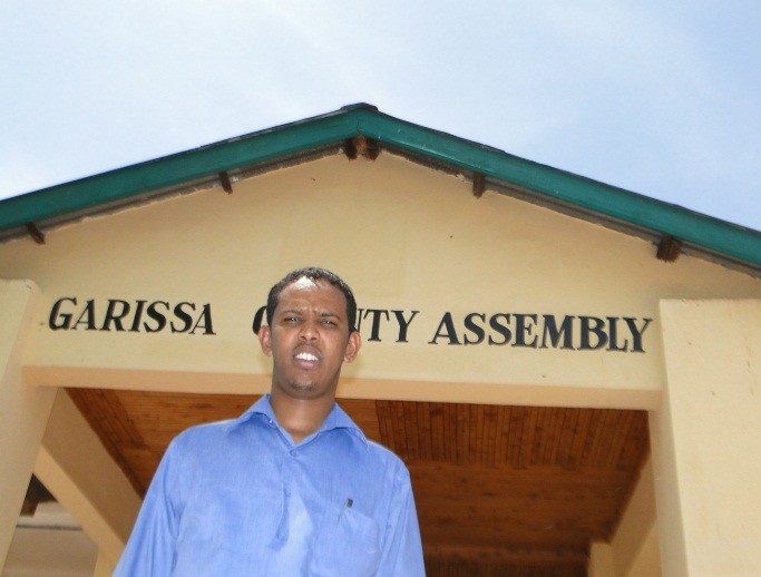 A young Kenyan man stands in front of a building labeled “Garissa County Assembly.”