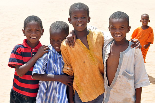 Four boys smile at the camera while a fifth boy looks on from the background
