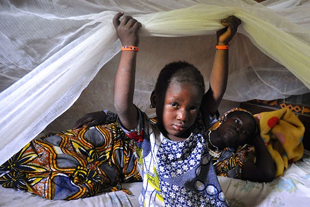 A young girl sits under a bednet with her mother behind her.