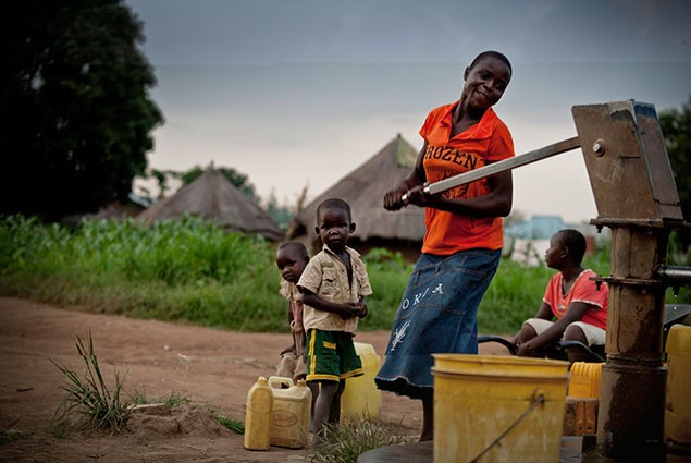 A woman pumps water into a bucket while children wait behind her to fill their containers