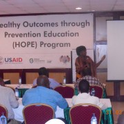 A Kenyan woman addresses an audience while standing in front of a banner that reads Health Outcomes Through Prevention Education